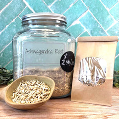 Glass jar, bowl, and paper bag labeled 'Ashwagandha Root' on a wooden surface with a tiled wall background. Sold by the ounce at Calhoun Farmstead.