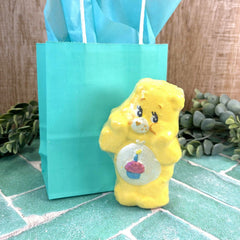 Yellow Care Bear figurine with a blue gift bag on a textured surface