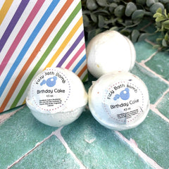 Two birthday cake scented bath bombs on a tiled surface with a striped bag and greenery in the background.