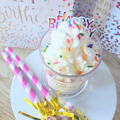 A decorative Happy Birthday dessert candle with a 'Happy Birthday' topper made to look like a dessert with whipped cream and sprinkles, accompanied by striped straws and a golden party blowout.