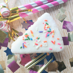 A cake-shaped bath bomb with white fondant-like topping and colorful sprinkles on a wooden surface surrounded by birthday decorations.
