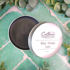Open tin of Calhoun Farmstead Black Drawing Salve on a green textured surface with eucalyptus leaves.