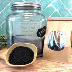 Glass jar with black seeds, wooden scoop, and brown paper bag on a wooden surface with a tiled wall background. Herbal teas sold at Calhoun Farmstead.