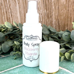 White body spray bottle labeled 'Blackberry Magnolia' by 'Cathleen Farmstead' on a green surface with greenery in the background. Natural body spray. Calhoun Farm self-care.