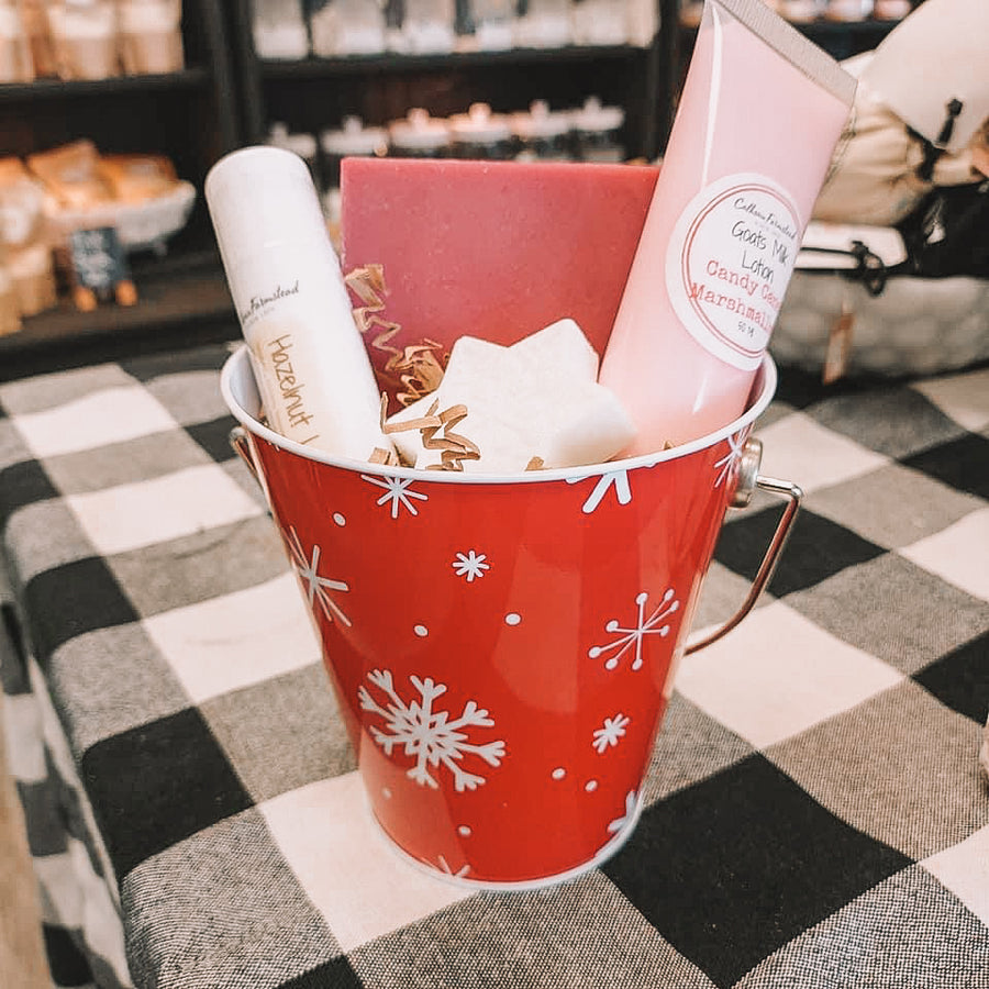 A Christmas gift set packaged in a milk bucket. Bucket is red with white snowflakes. Contents of pail include goats milk lotion tube, jumbo lip balm, goats milk soap, and a snowflake shaped guest soap.