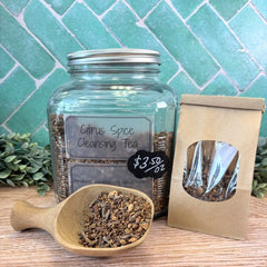 Jar of Citrus Spice Cleansing Tea with a scoop and packaging on a wooden surface with a green tiled background. Herbs for cleansing.