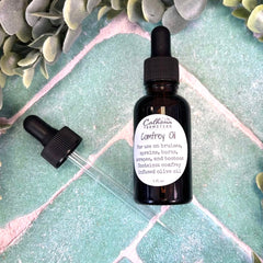 Bottle of Calhoun Farmstead Comfrey Oil with dropper on a green tiled surface with leaves.