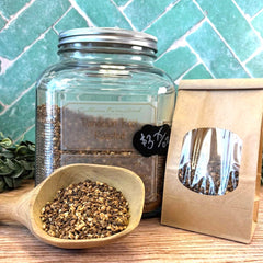Glass jar with roasted dandelion root, wooden scoop, and packaging on a wooden surface with a green tiled wall background. Dandelion root tea. Roasted dandelion.
