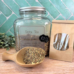 Glass jar of Detox Tea with a wooden scoop and packaging on a wooden surface with a green tiled wall background. Herbs for detoxing. Teas for detox.