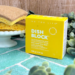 Yellow box of 'Dish Block' soap on a tiled surface with sponges and plants in the background. Solid Dish Soap