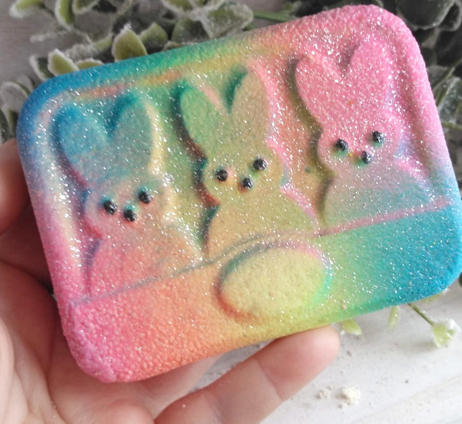 Hand holding a bath bomb decorated with three Easter peep bunny shapes. Colored with light pink, blue and yellow airbrushing.