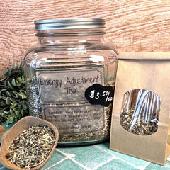 Glass jar labeled 'Energy Adjustment Tea' with a small bag of tea leaves on a wooden surface. Caffeine free tea. Energy tea.