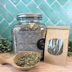 Glass jar and paper bag of 'Feminine Tea Blend' with a wooden scoop on a wooden surface. Feminine Tea Blend. Hormone support. Herbs for menopause.