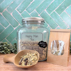 Glass jar of 'Happy Man Tea' with a wooden scoop and packaging on a wooden surface with a green tiled wall background. Teas for men. Herbs for men. Mens Herbal wellness.