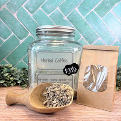 Glass jar labeled 'Herbal Coffee' with a wooden scoop containing coffee grounds on a wooden surface with a green tiled wall background. Dandelion tea. Herbal coffee.