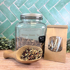 Tea jar, scoop with tea leaves, and packaging on a wooden surface with a tiled wall background. Hibiscus Heaven Tea. Floral teas.