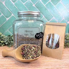 Glass jar with hibiscus high tea, wooden scoop, and packaging on a wooden surface with a green tiled background. Hibiscus Teas. Floral tea. Honeybush tea.
