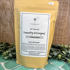 Yellow package of Dandelion Naturals All Natural Laundry Detergent on a wooden background with greenery.
