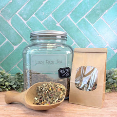 Glass jar labeled 'Lazy Daze Tea' with a wooden scoop and tea bag on a wooden surface with a teal tiled wall background. Herbal teas in Pennsylvania.