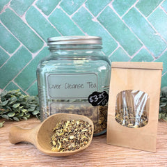 Glass jar labeled 'Liver Cleanse Tea' with a wooden scoop and packaging on a tiled background. Herbs for detox. Metal detox.