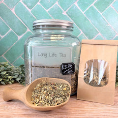 Glass jar of 'Long Life Tea' with a wooden scoop and tea package on a wooden surface with a tiled wall background. Herbs for overall wellness. Herbal apothecary.