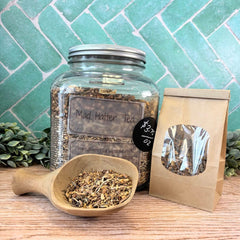 Glass jar of 'Mad Hatter Tea' with a wooden scoop and tea package on a wooden surface with a green tiled wall background. Fun teas for parties. Drinking tea.