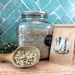 Glass jar with dried herbs, wooden scoop, and packaging on a tiled wall background. Herbs for cough, sore throat, and colds.