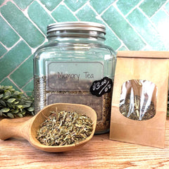 Glass jar labeled 'Memory Tea' with a wooden scoop and packaging on a tiled background. Herbs for memory and brain health.