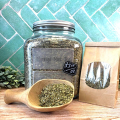 Glass jar with Moringa Mint Tea, wooden scoop with tea leaves, and brown paper bag on a wooden surface with green tiled wall background. Benefits of moringa.