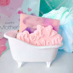 A small plastic bathtub soap dish with a 'Happy Mother's Day' card, containing a pink bar of goat's milk soap with 'MOM' written on it, against a floral patterned background.