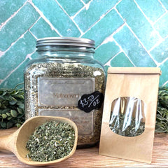 Glass jar with silver lid, wooden scoop with herbs, and brown paper bag on a tiled background. Motherwort herb for women's health. Herbs for women.