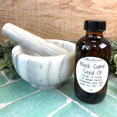 Brown glass bottle of Black Cumin Seed Oil with a label next to a marble mortar and pestle on a tiled surface.