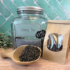 Oolong tea in a glass jar, wooden scoop, and brown paper bag on a wooden surface with a green tiled background. Oolong black tea.