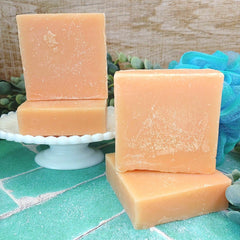 Three bars of soap on a white stand with a blue loofah and greenery in the background.