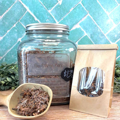 Clear jar with labeled tea leaves, small scoop of tea leaves, and brown paper bag on a wooden surface with a tiled wall background. Pau D'Arco Bark. Herbal remedies for illness.