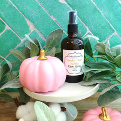 An amber glass room spray bottle labeled 'Pumpkin Spice Waffles Room Spray' by Colleen Reifallou, styled on a white pedestal with a pink decorative pumpkin, green foliage, and a green tile background.