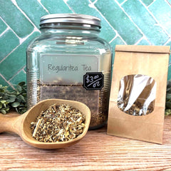 Glass jar of tea with a wooden scoop and packaging on a wooden surface with a teal tiled background. Digestion tea. Herbs for digestion.
