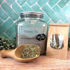 Glass jar of 'Relaxing Tea' with a wooden scoop and tea bags on a wooden surface with a teal tiled wall background. Teas for relaxation. Herbs for sleep.