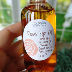A hand holding a 2oz glass dropper bottle of Rose Hip Oil with a label that includes the product name and a list of ingredients such as Rose Hips, Squalene Oil, Tamanu Oil, Sweet Almond Oil, and Rose Essential Oil.