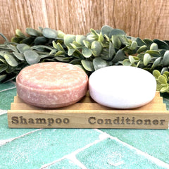 Two soap bars on a wooden stand labeled 'Shampoo' and 'Conditioner' with greenery in the background. Solid shampoo and conditioner bar holder. NO plastic. Sustainable.
