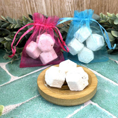 White shower steamers on a wooden plate with pink and blue drawstring bags in the background. Mini shower steamers. Relaxing shower. 