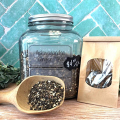 Jar of Ashwagandha Ginger Tea with a wooden scoop and packaging on a tiled background