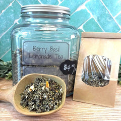 Glass jar of Berry Basil Lemonade Tea with a wooden scoop and tea bags on a wooden surface with a blue tiled wall background.