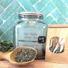 Glass jar of Gun Powder Green Tea with a wooden scoop and packaging on a tiled background. Camelia sinensis tea. Green Tea. 