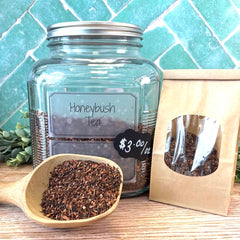 Glass jar of Honeybush Tea with a wooden scoop and packaging on a teal tiled background