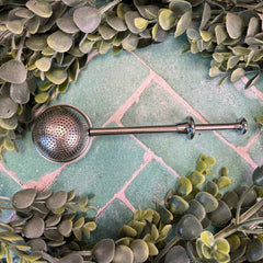 Metallic tea infuser on a green tiled surface with greenery around. Tea accessories. Loose leaf tea infuser