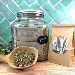 Glass jar of Razzlemint Tea with a wooden scoop and paper bag on a tiled background