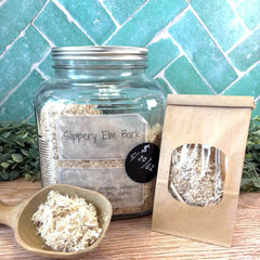 Glass jar with slippery elm bark, a wooden scoop, and a paper bag on a wooden surface with a tiled wall background. Herbal apothecary.