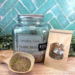 Glass jar labeled 'Sprouting Seeds' with a wooden scoop and paper bag on a wooden surface. Sprouting seeds. Gardening seeds. Sprouts.