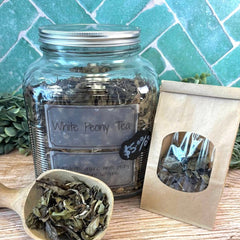 Glass jar of White Peony Tea with a scoop and packaging on a wooden surface with a tiled background.